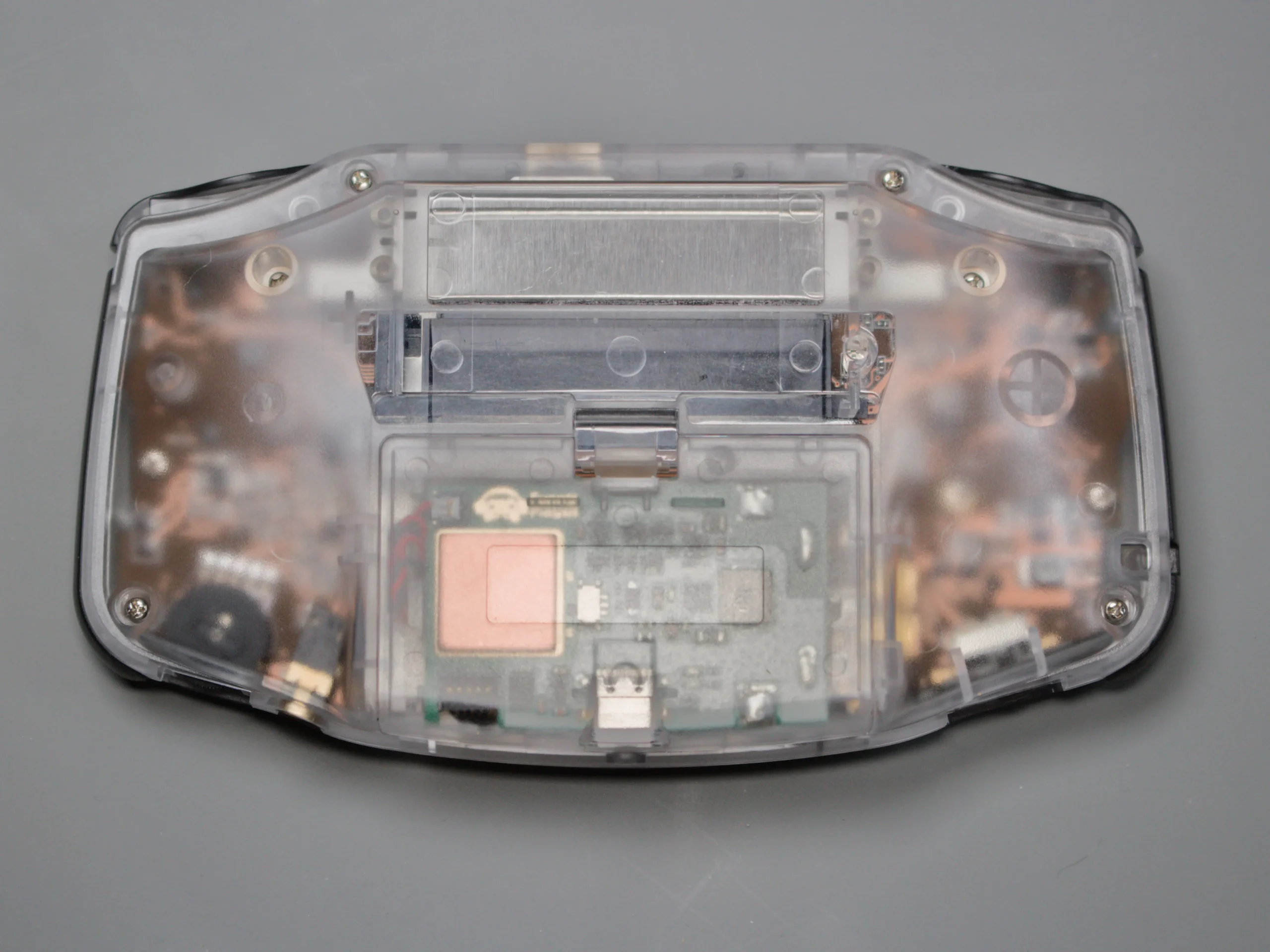 Rear view of a clear-shell Game Boy Advance showing the battery compartment and internal electronics through the case