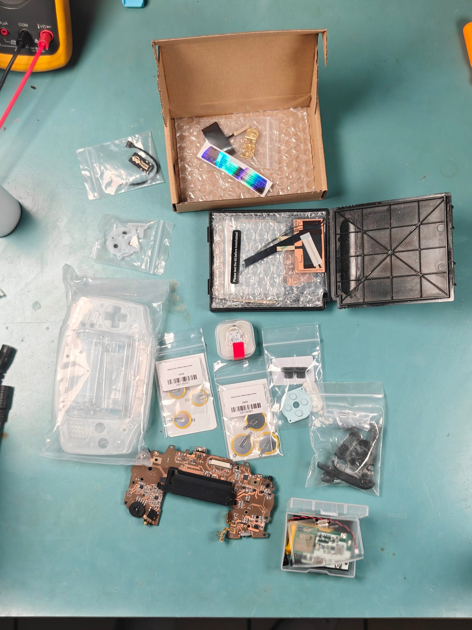 Parts gathered for a Game Boy Advance rebuild, including the motherboard, clear shell, replacement screen, buttons, batteries, and small components